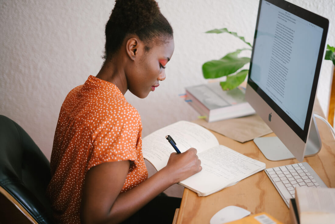 A woman writing in a notebook in front of a desktop computer